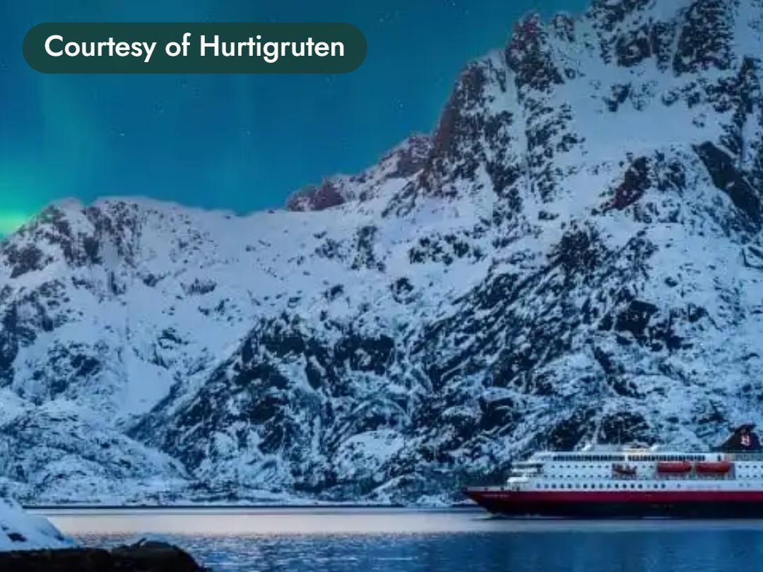 A ship sails below snowy mountains under the northern lights in Norway. Courtesy of Hurtigruten.