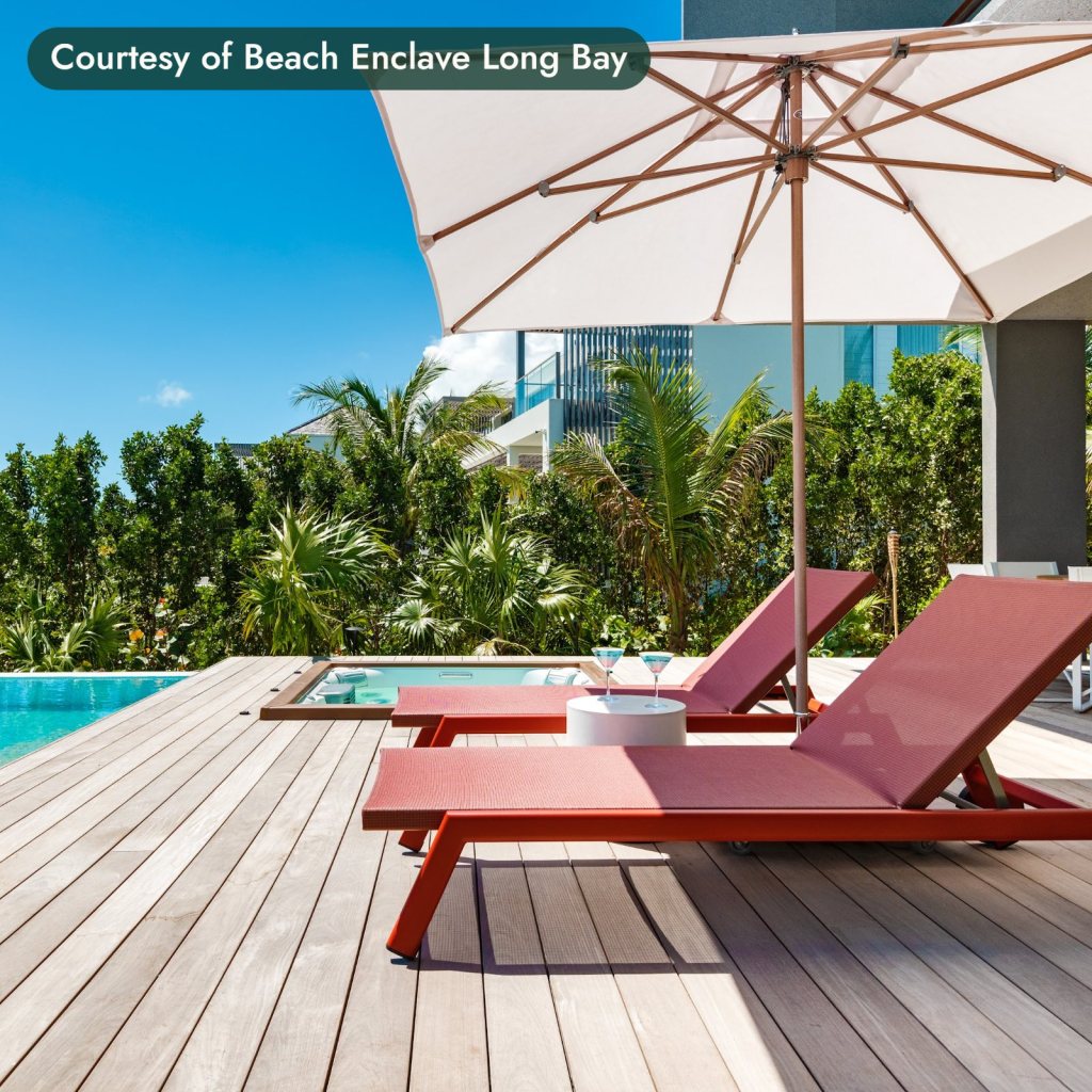 Red loungers under a white umbrella on a pool deck framed by palms in Turks and Caicos. Courtesy of Beach Enclave Long Bay.