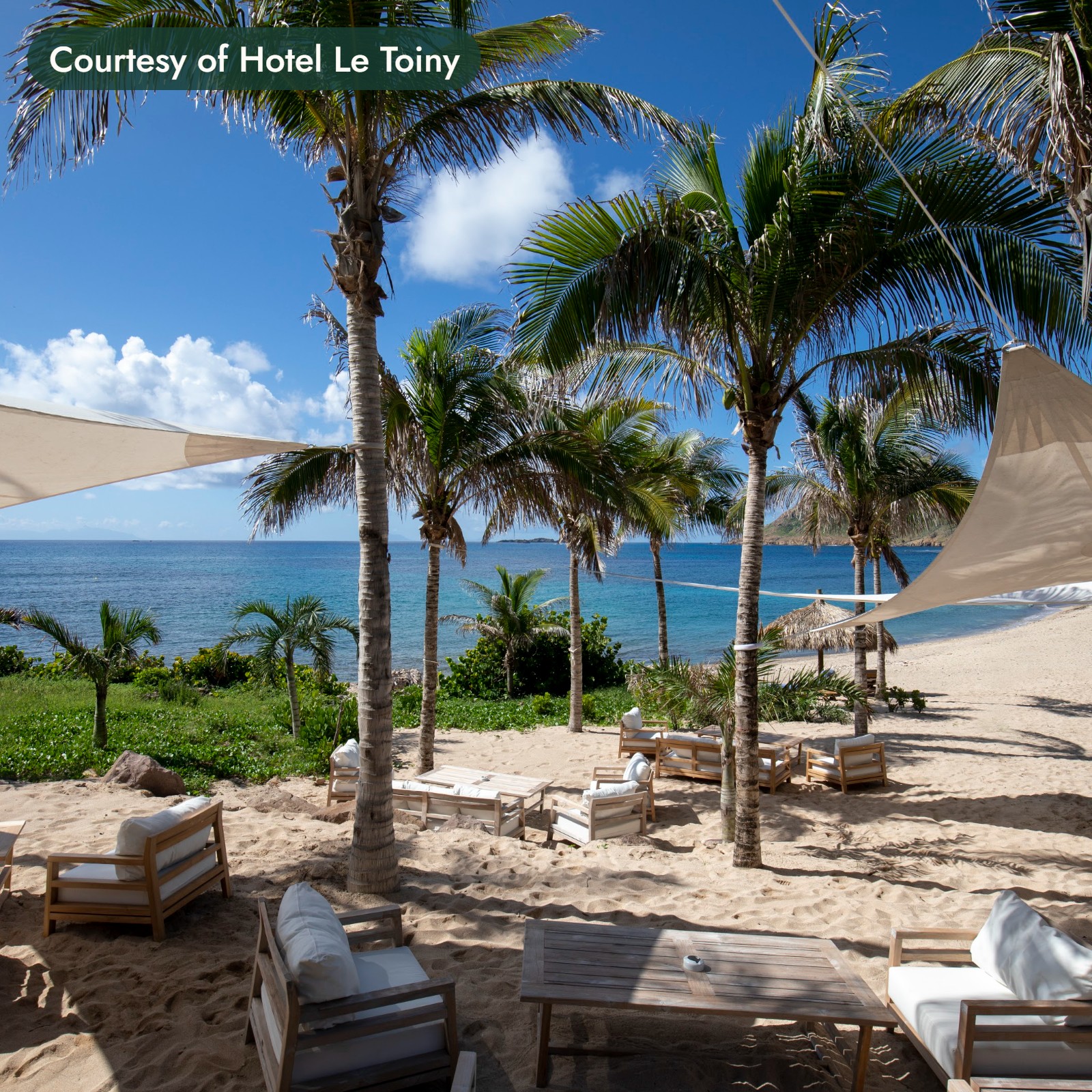 Teak lounge chairs under sail shades and palm trees on a beach in St. Barths, with the sea beyond. Courtesy of Hotel Le Toiny.