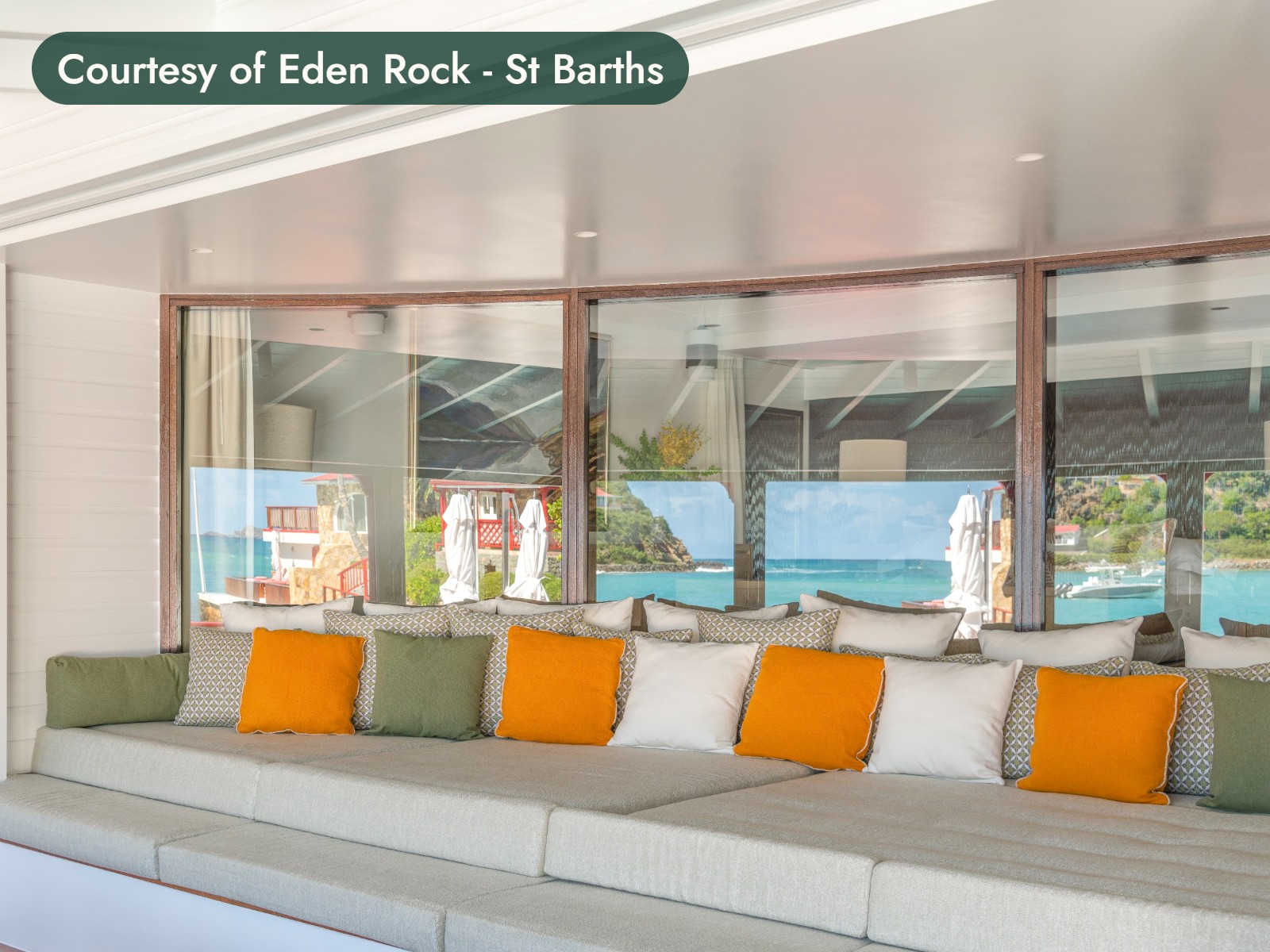 A long banquette with orange and green cushions in a suite lounge, its windows looking out to the sea in St. Barths. Courtesy of Eden Rock - St Barths.