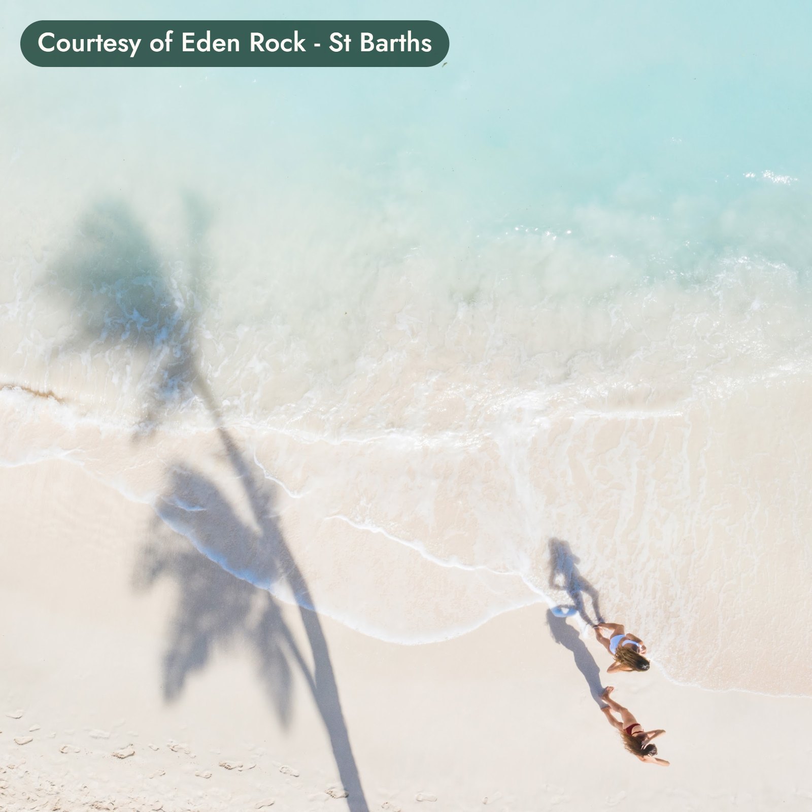 Two travelers walk along the water’s edge in St. Barths beside the long shadow of a palm. Courtesy of Eden Rock - St Barths.
